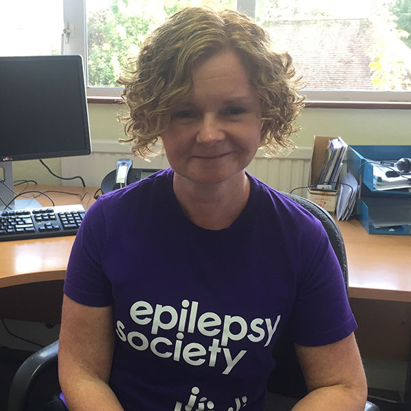 Andree Mayne has a blonde, curly bob. She is wearing a purple Epilepsy Society t-shirt and is smiling.