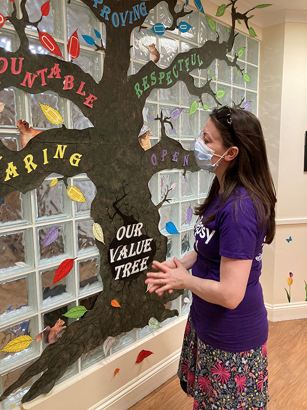 Sarah is wearing a purple Epilepsy Society t-shirt and standing in front of a values tree in one of the residential care homes.