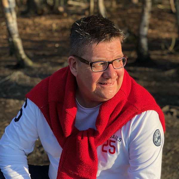 ​​​​Steve Turner is wearing a whilte sports shirt with a red jumper tied around his shoulders. He has spiky hair, glasses, a friendly smile and is looking off camera, over his right shoulder