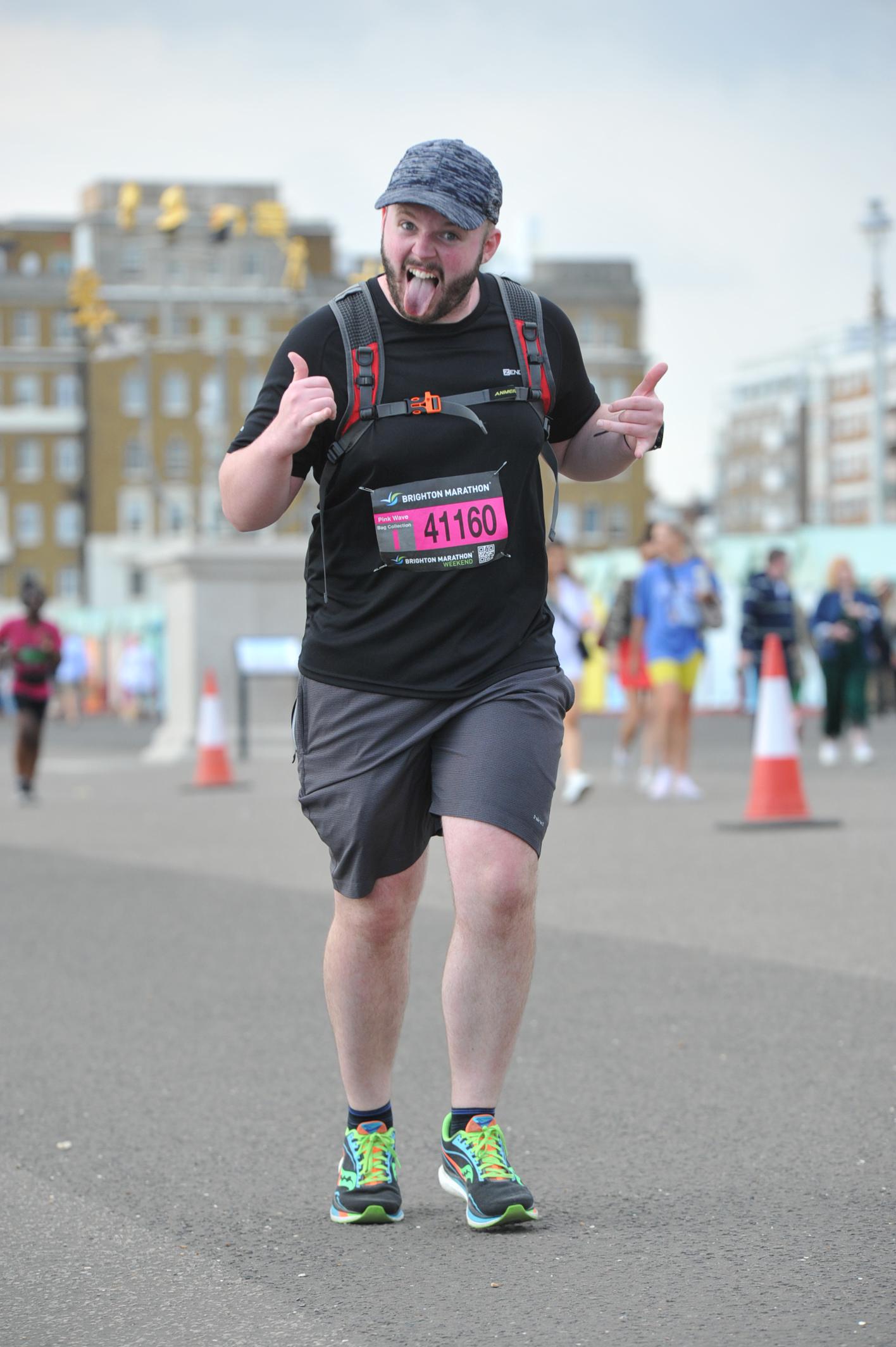 Alex, AKA The Fifa Analyst, running in the Brighton Marathon on pavement, with his thumbs up and poking his tongue out to the camera, wearing a baseball cap, grey shorts, black t-shirt and neon trainers